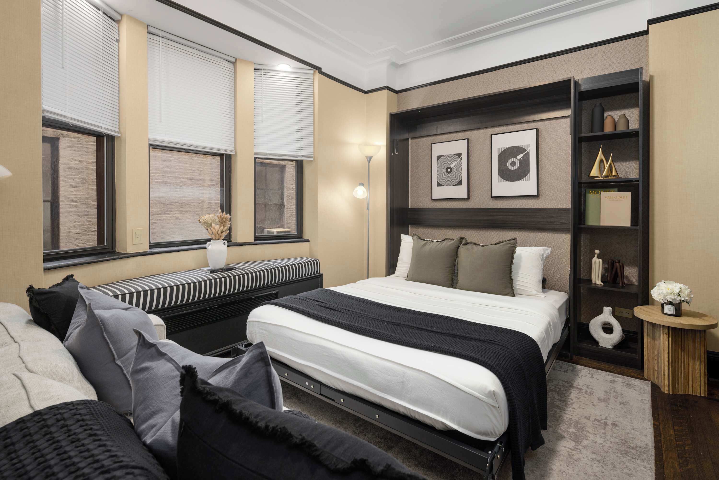 Ein modernes Schlafzimmer mit einem großen Bett, einem Nachttisch, einem Bücherregal und einem Fenster mit Jalousien.