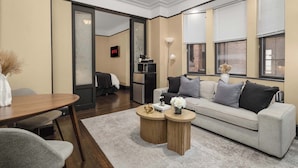 A modern living room with a sofa, coffee table, and a dining table. There is a TV mounted on the wall and a lamp on a side table. - Premier Midtown Stay | Walk to Times Square! (New York)