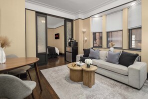 A modern living room with a sofa, coffee table, and a dining table. There is a TV mounted on the wall and a lamp on a side table. - Premier Midtown Stay | Walk to Times Square! (New York)