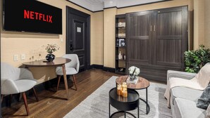 Un salon moderne avec un téléviseur à écran plat affichant le logo « NETFLIX », une armoire en bois foncé, une table basse ronde ornée de bougies et un canapé gris.