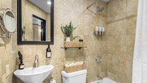 Baño con lavamanos, inodoro y bañera. Incluye un espejo, repisas con artículos de aseo personal y zona de ducha.