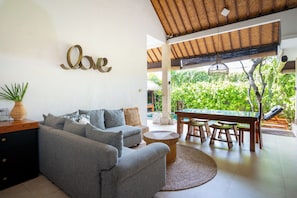 A modern living room with a grey sofa, a wooden coffee table, and a dining area with wooden stools. A large 'love' sign is on the wall, and there is a view of lush greenery outside. - 100m to Seminyak Beach / Personal Full-Time Butler (Seminyak)
