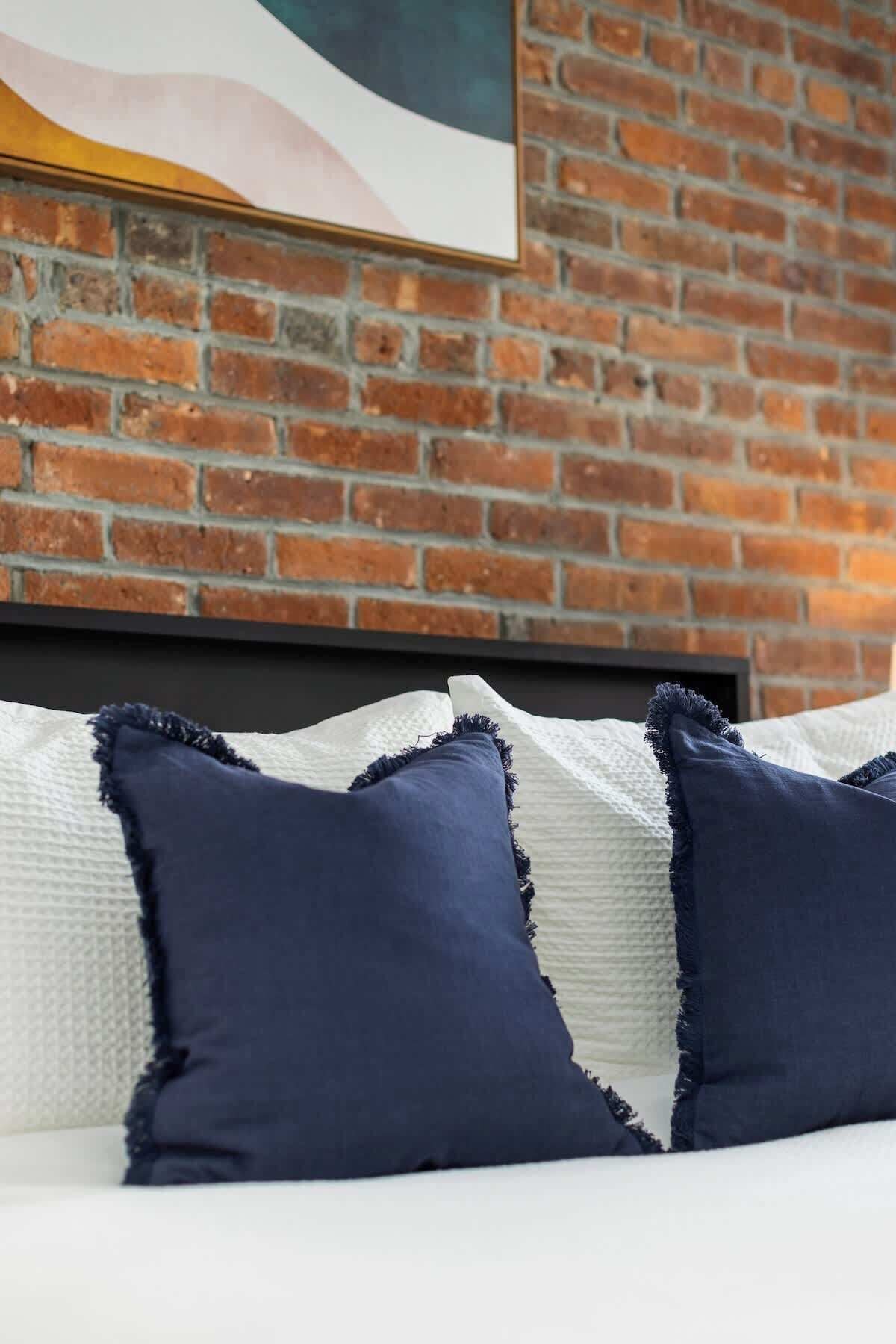A bed with white and dark blue pillows against a brick wall.