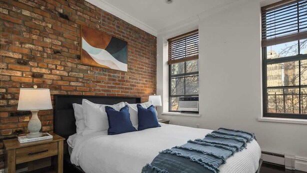 A bedroom with a brick wall, a large window, a bed with white and blue bedding, and a nightstand with a lamp. - Chic 2BR Retreat in the Heart of Times Square (New York)