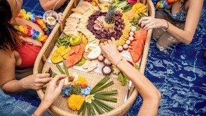 Un grupo de personas alrededor de una bandeja grande de madera repleta de frutas, verduras y bocadillos.