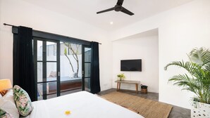 Habitación de hotel moderna con una cama grande, ventilador de techo, televisión de pantalla plana, una pequeña mesa con una planta y vistas a través de una puerta corrediza.