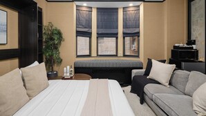 Quarto de hotel com uma cama grande, um sofá, uma mesinha, um vaso de plantas e um parapeito com cortinas.