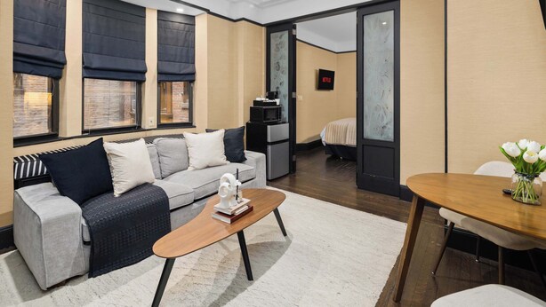 Quarto de hotel moderno com sofá, mesa de centro de madeira, mesa de jantar com cadeiras e televisão na parede.