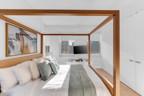 A modern bedroom with a wooden canopy bed, a large painting, a flat-screen TV, and a city view.
