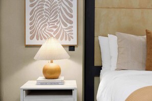 Un lit bien fait, avec des oreillers blancs et bruns, une table de chevet sur laquelle se trouvent une lampe et un livre intitulé « Améliorez et décorez », et une œuvre d’art représentant une feuille, encadrée, accrochée au mur.