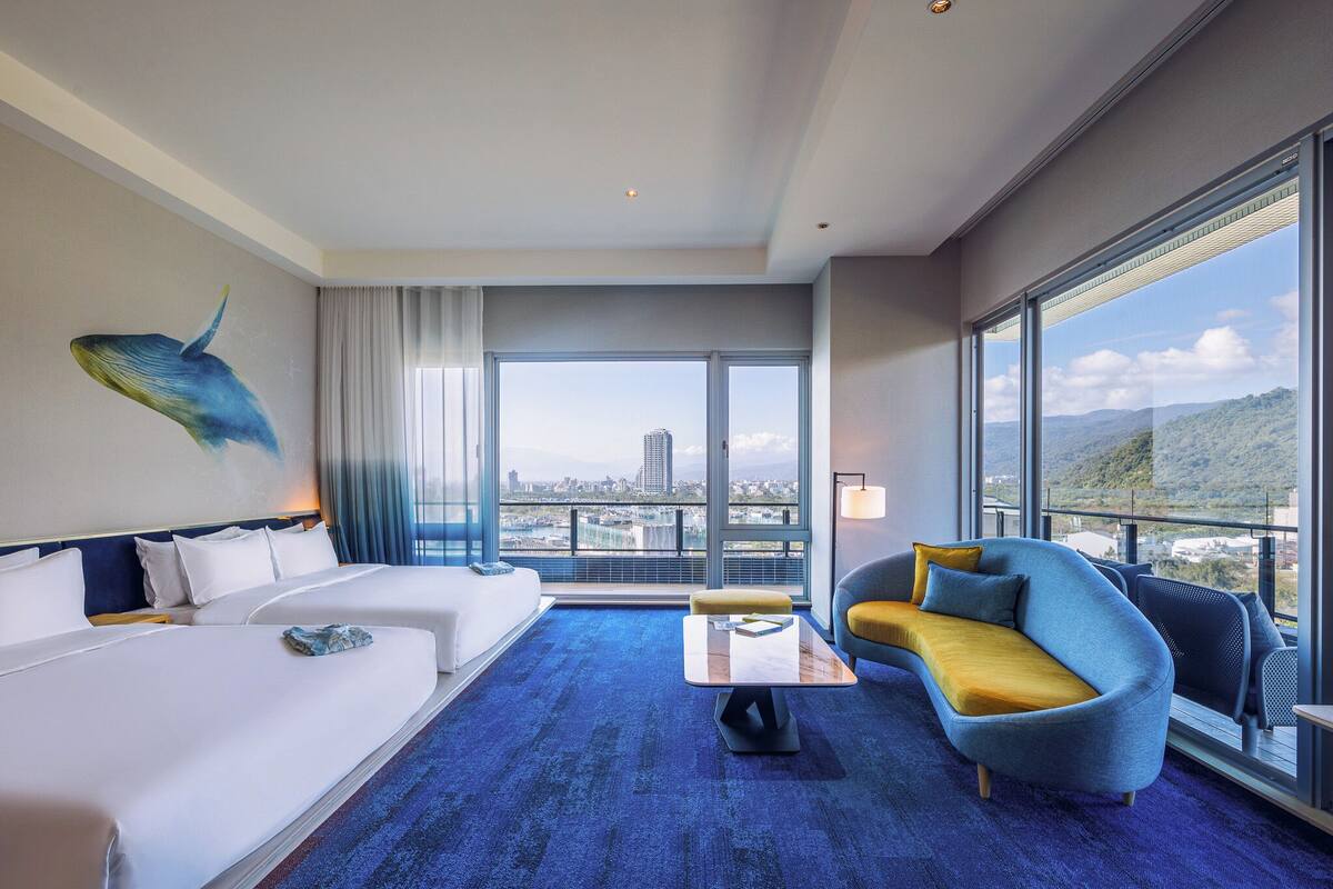 A hotel room with a large bed, a sofa, a coffee table, and a view of the cityscape through large windows.