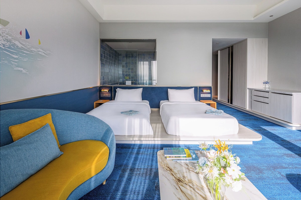A modern hotel room with a blue and yellow sofa, two single beds, a marble coffee table with flowers, and a view of the bathroom.