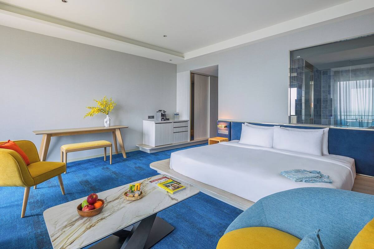 A hotel room with a large bed, a desk, a chair, a coffee machine, and a view of the city through the window.