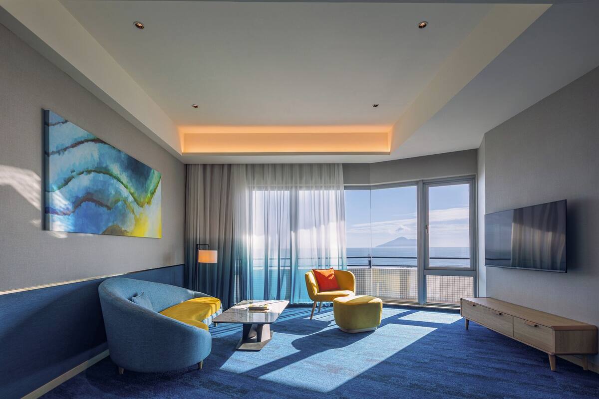 A modern hotel room with a large window, a flat-screen TV, a sofa, a chair, and a coffee table.