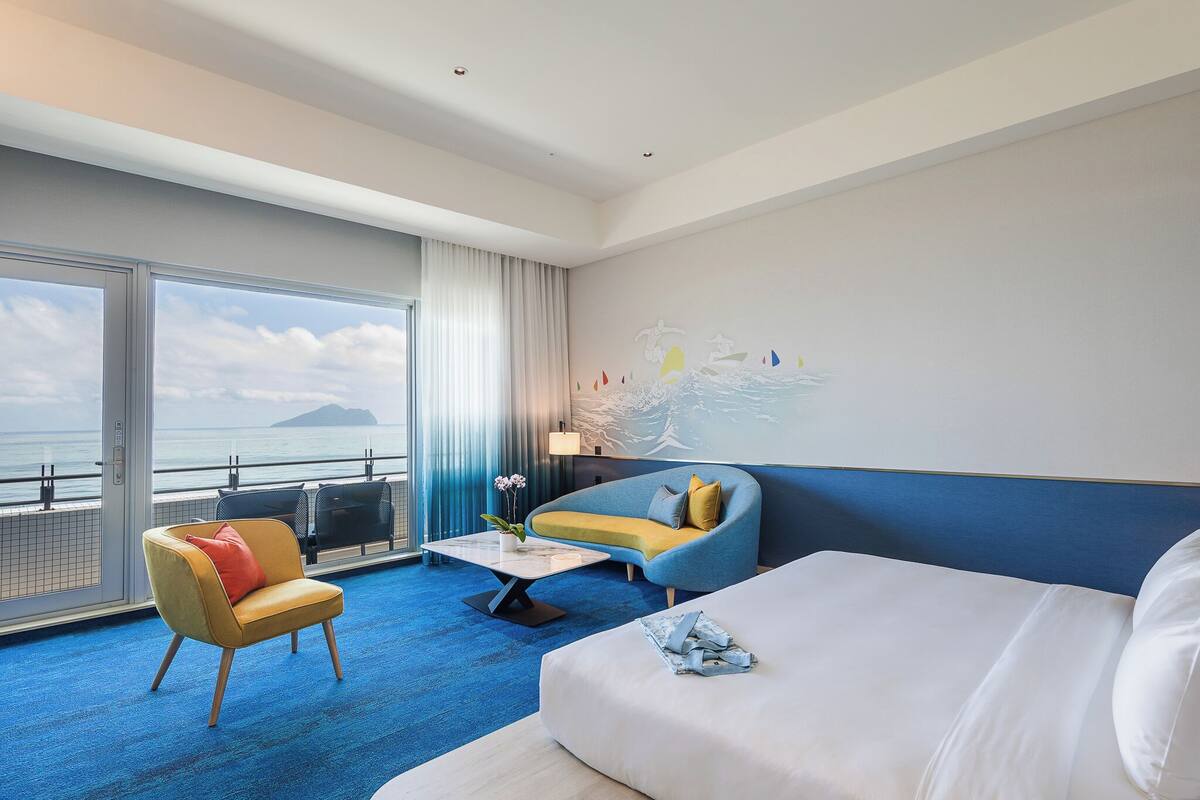 A modern hotel room with a large bed, a sofa, a chair, and a view of the sea.