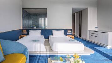 Habitación de hotel con dos camas, un sofá, una mesita con libros y flores, y un baño visible a través de una puerta de cristal.