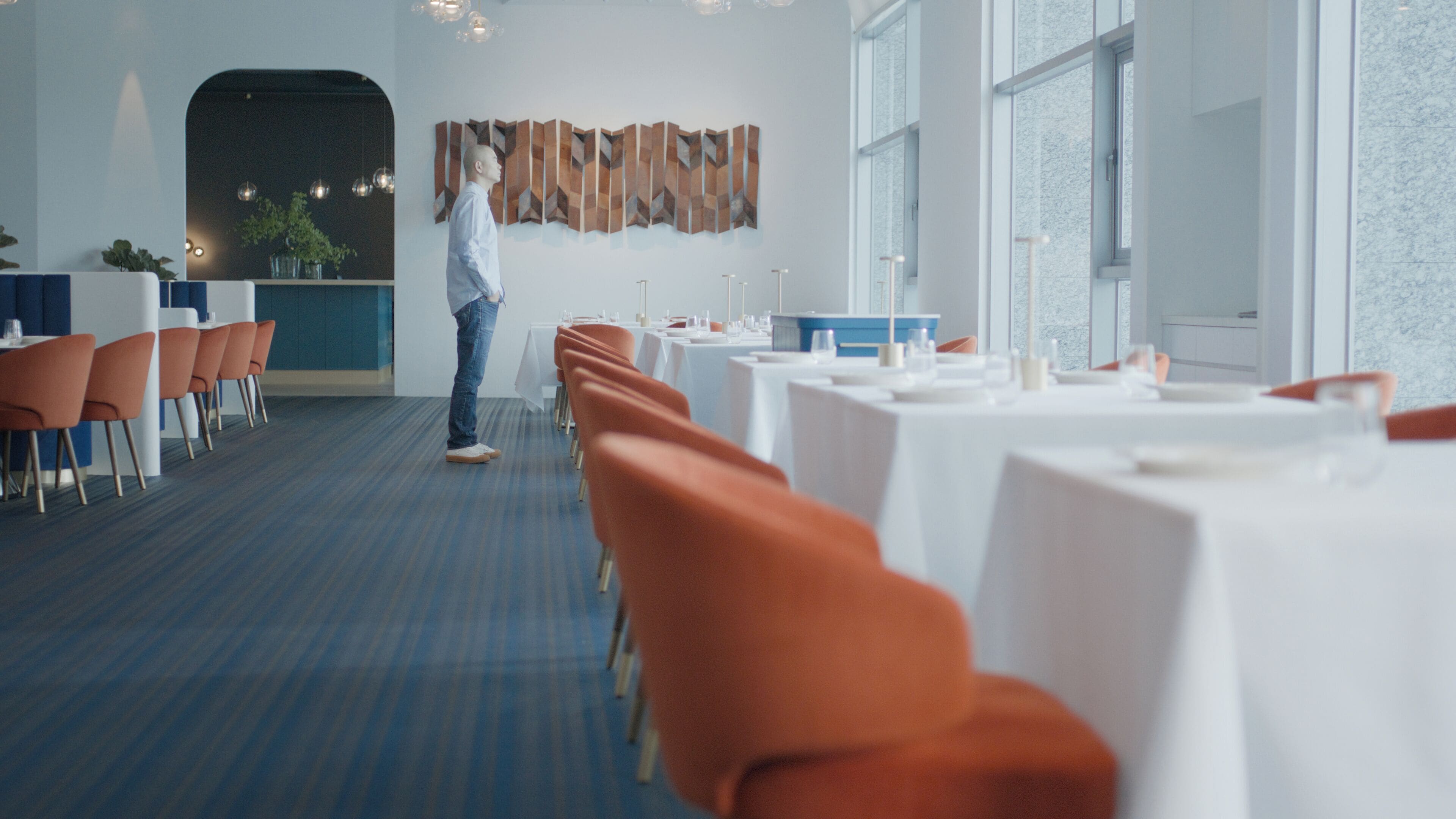 A modern dining area with a long table, orange chairs, and a person standing.