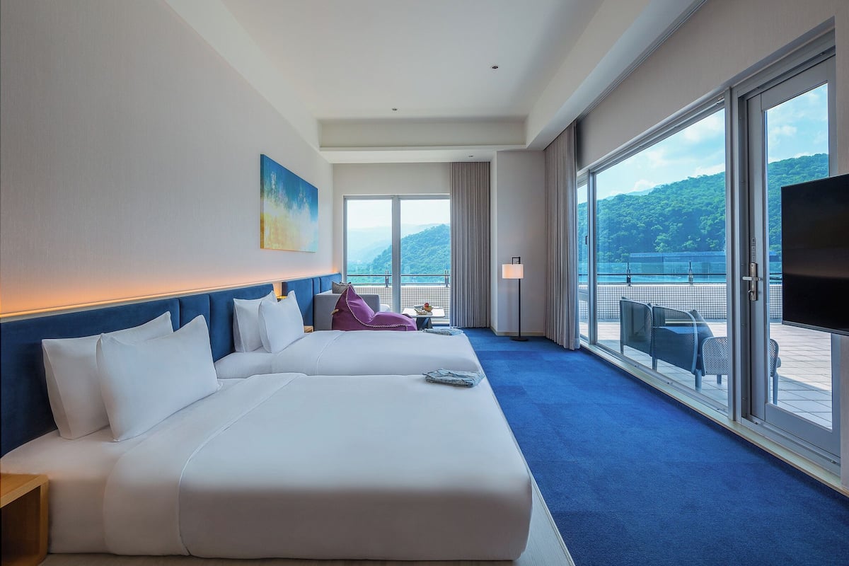 A hotel room with two beds, a large window with a view of a mountain and water, and a balcony with seating.