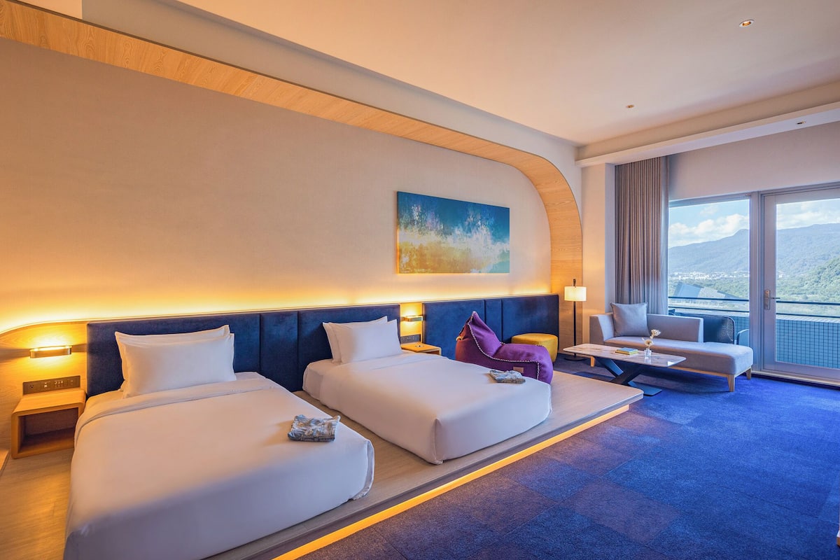 A hotel room with two beds, a sofa, a small table, and a large window offering a city view.