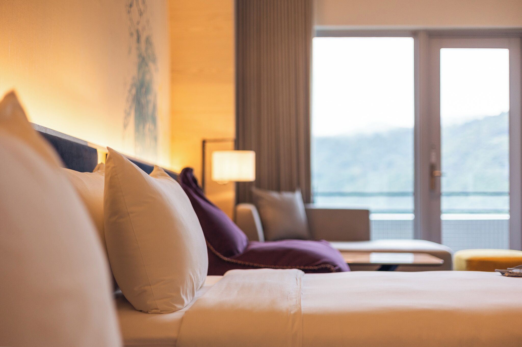 A hotel room with a bed, pillows, a lamp, and a view of the outdoors.