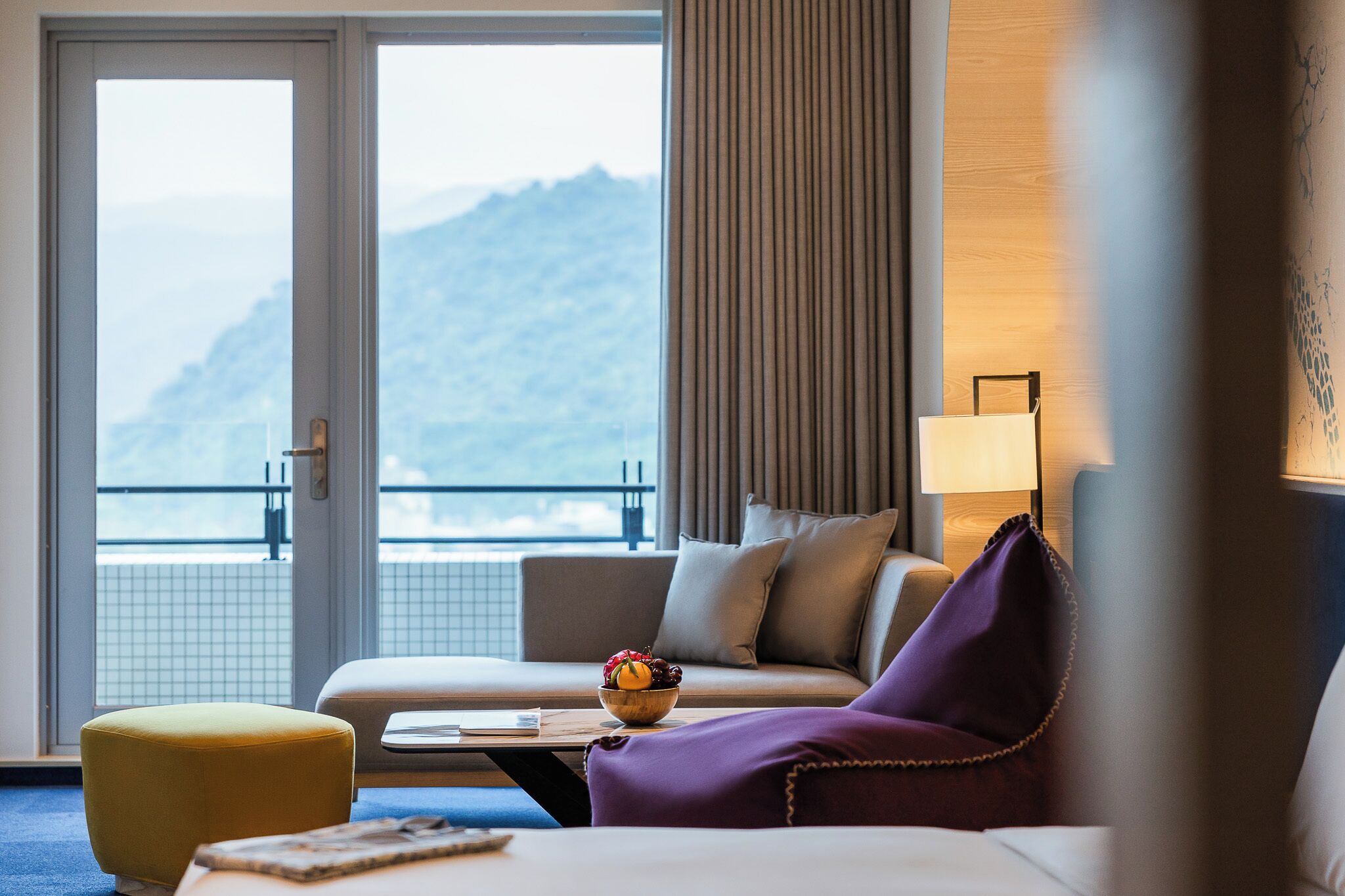 Habitación de hotel con sofá, una mesita con un cuenco de fruta, un sillón morado y vistas a las montañas a través de una puerta corredera.