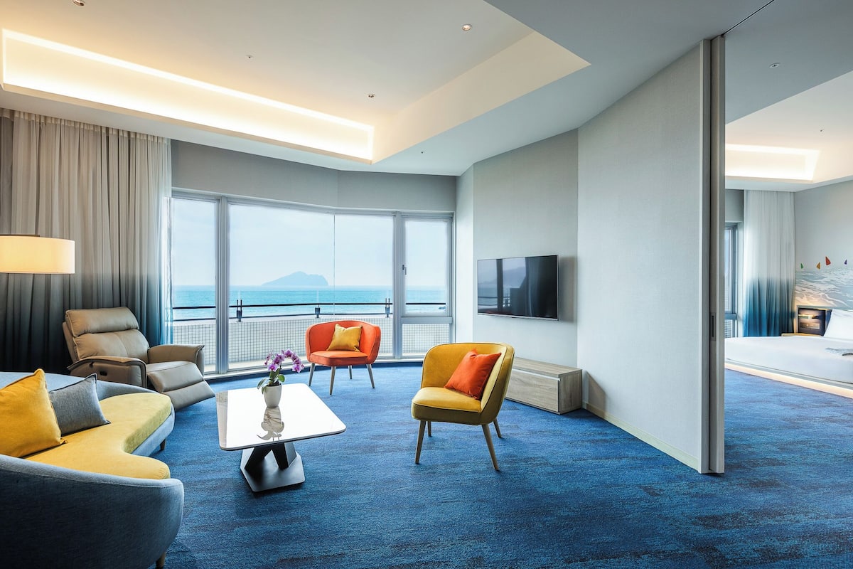 A modern hotel room with a large window, a flat-screen TV, and a cozy seating area with a sofa and armchairs.