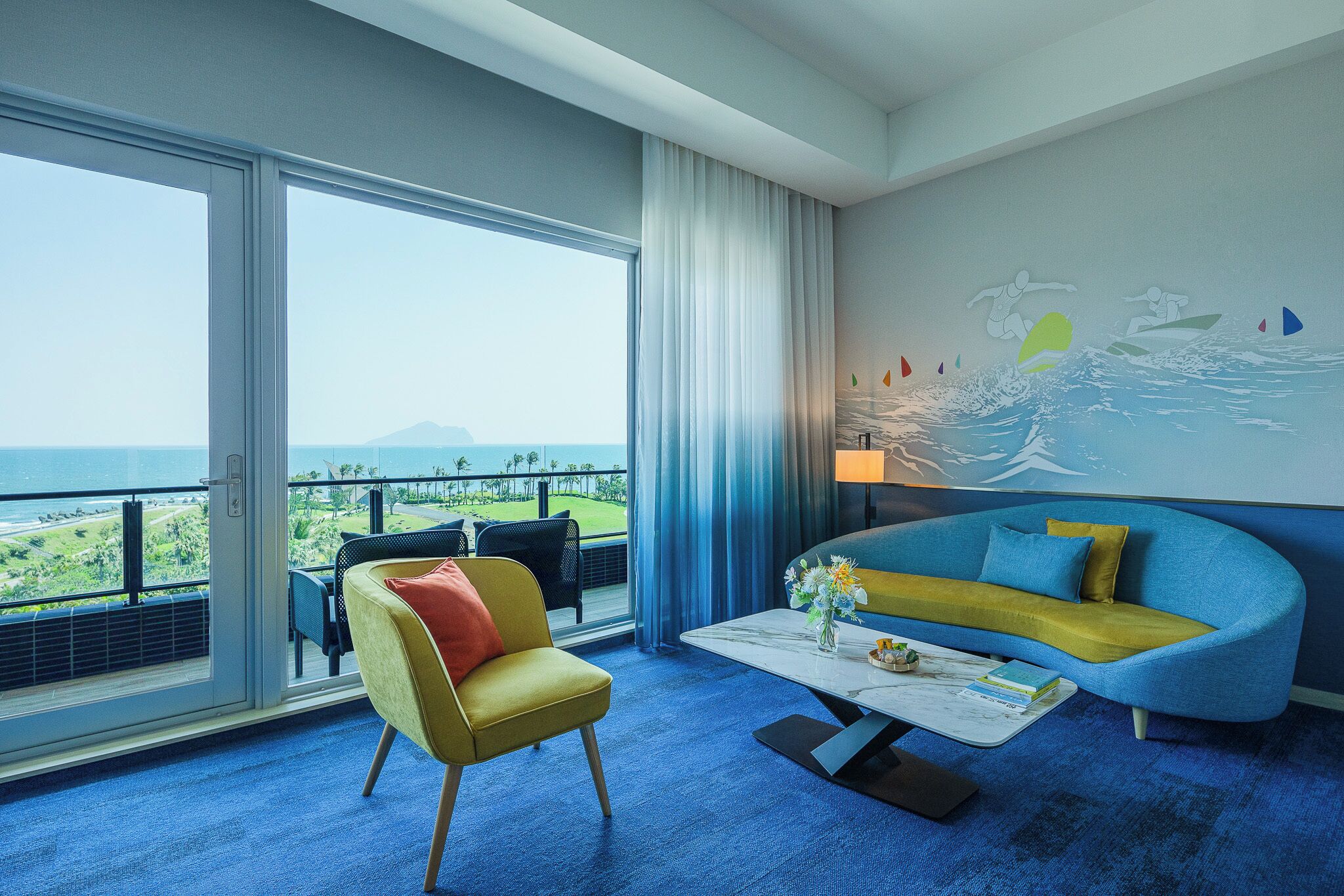 Una habitación de hotel con un ventanal grande, un sofá azul, un sillón amarillo y un mural de surfistas en la pared.