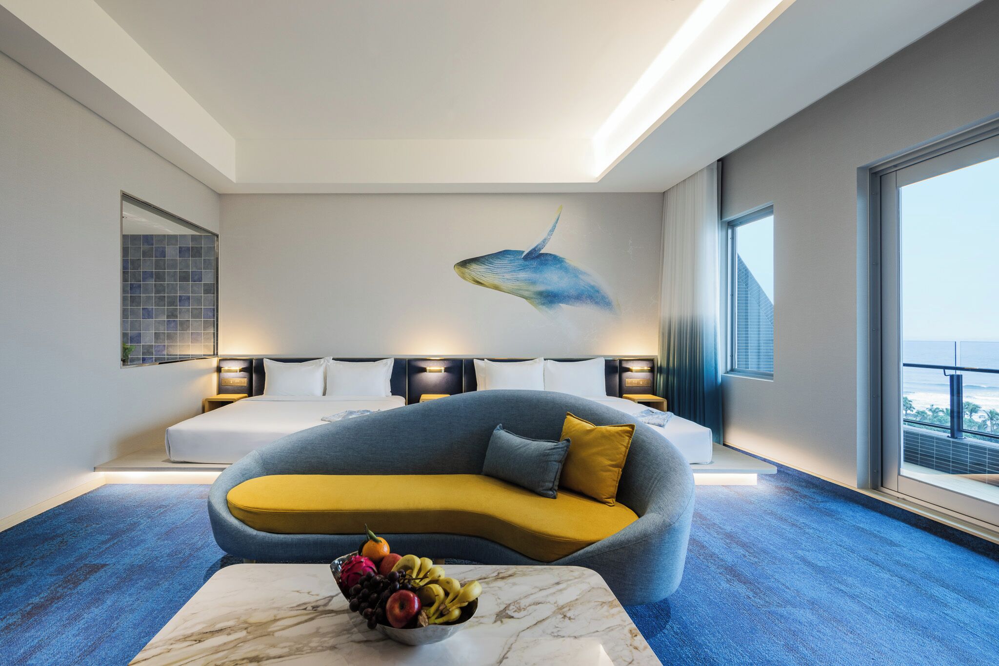 A modern hotel room with a curved sofa, a marble coffee table with fruit, a wall mural of a whale, and a large window offering a city view.