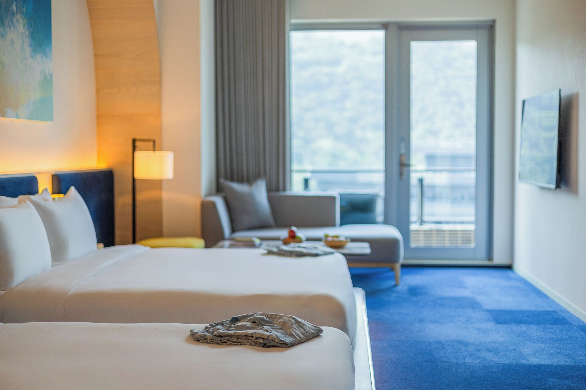 A hotel room with a large bed, a sofa, a small table, and a balcony with a view.