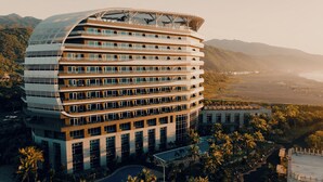 Un hotel de varios pisos con fachada curva, balcones y un área de piscina rodeada de palmeras.