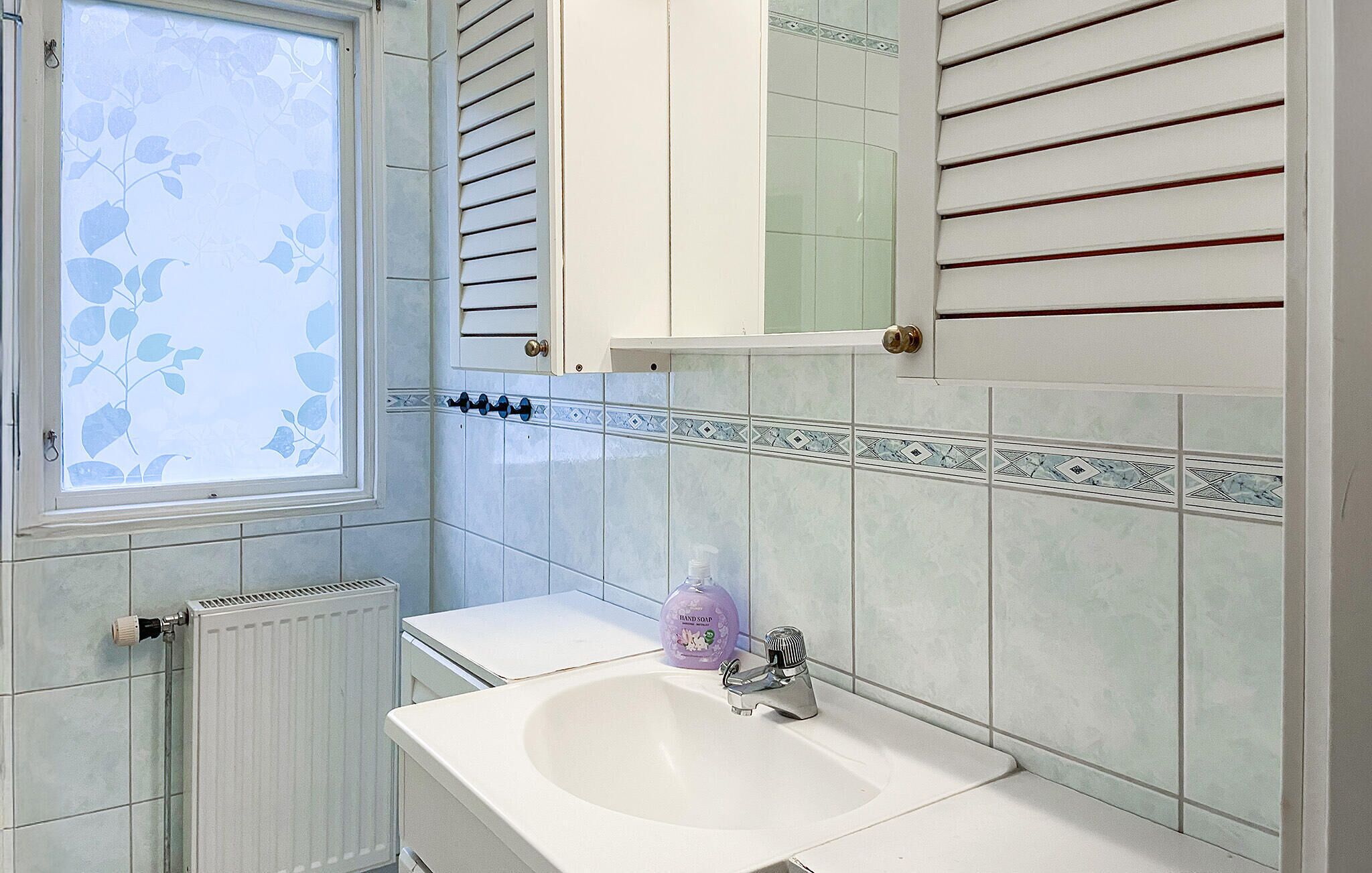 A bathroom with a window, radiator, sink, and hand soap dispenser.