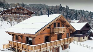 Una amplia cabaña de madera con varios balcones y un techo cubierto de nieve, rodeada de un paisaje montañoso boscoso.