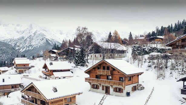 Un pueblo cubierto de nieve con casas de madera, una cabaña de madera y montañas al fondo.