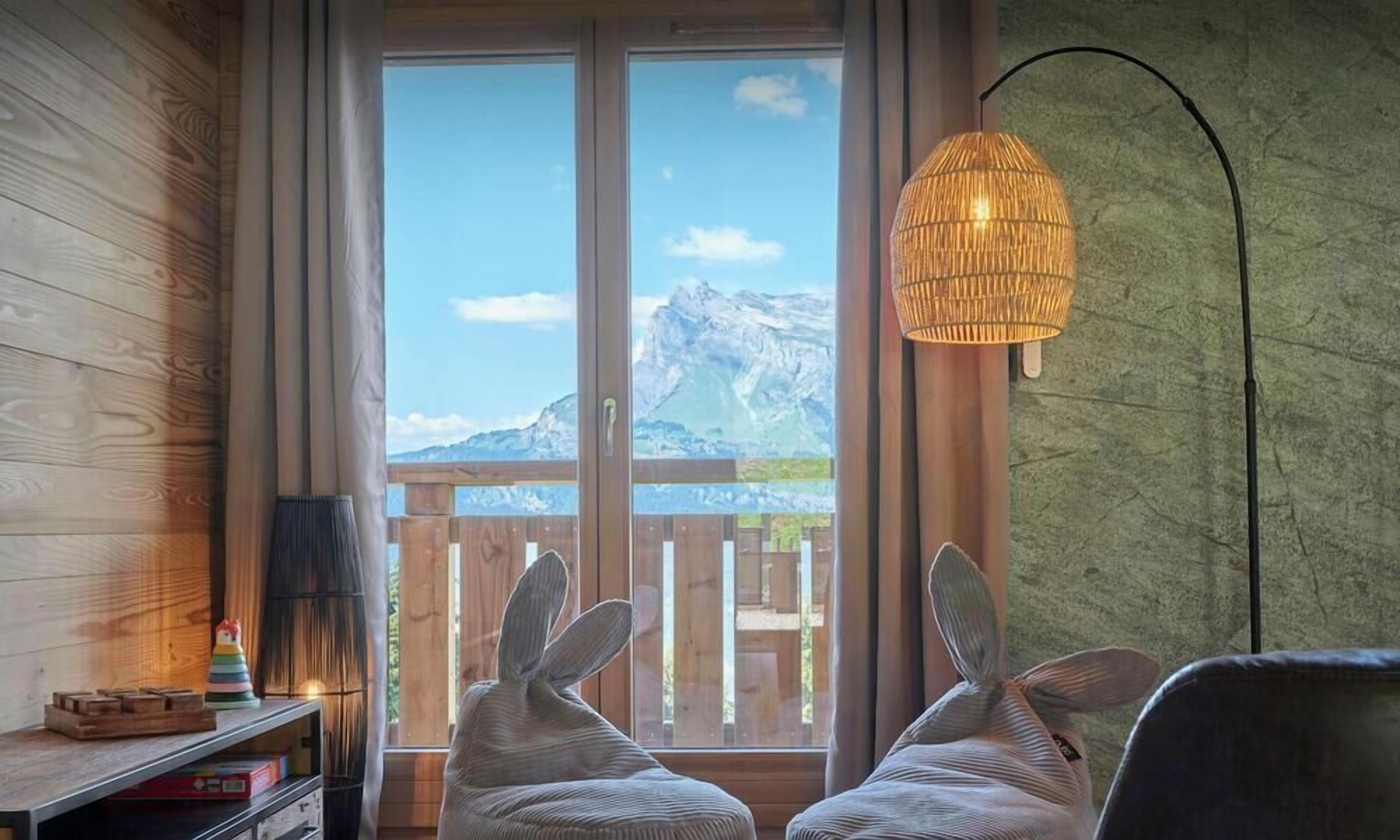 Ein Zimmer mit Holzboden, einem Fenster mit Bergblick, einem Korbstuhl und einer Pendelleuchte.