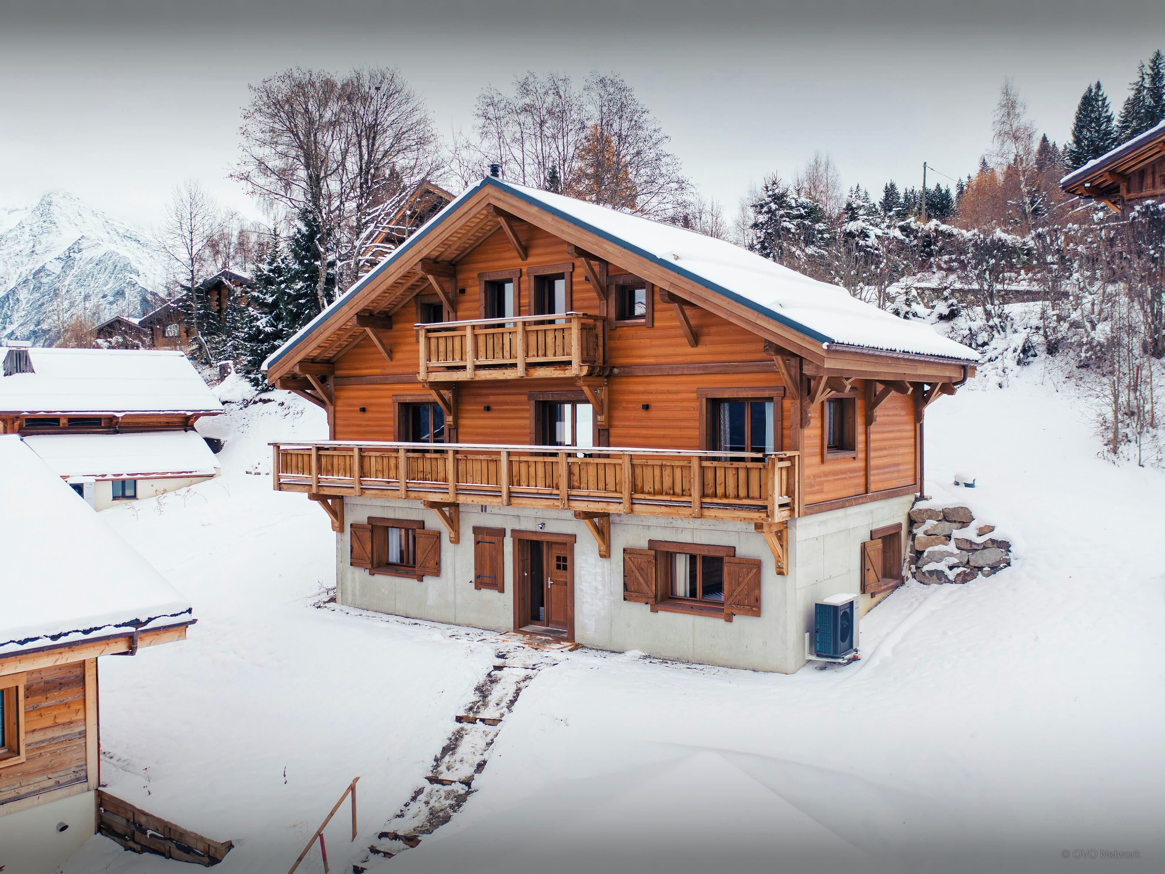 Un chalet en bois à plusieurs étages, doté d’un balcon, entouré d’arbres couverts de neige et avec une montagne en arrière-plan.