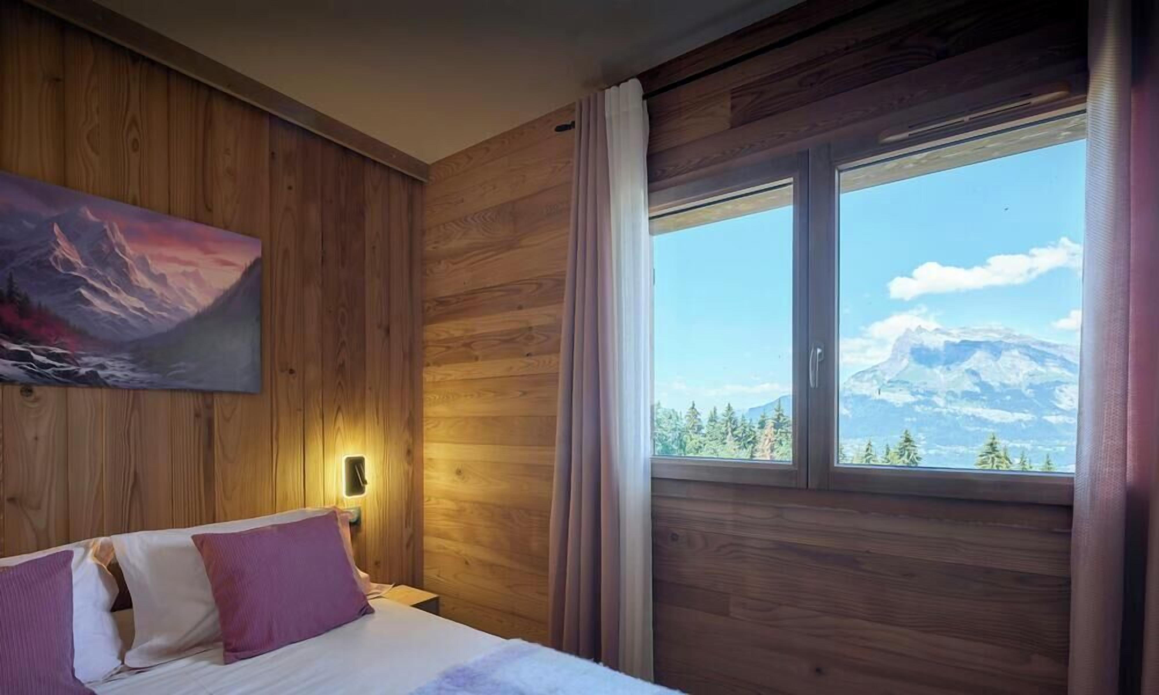 A wooden room with a bed, a painting, a window with a mountain view, and a nightlight.