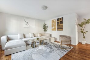 Un salon moderne avec un canapé d’angle, une table basse en verre et un tapis en marbre. On y trouve deux œuvres d’art murales et une plante en pot.