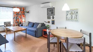 Uma sala de estar com sofá, mesa de jantar, cadeiras, mesa de centro, abajur e janela com cortinas.