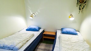 Quarto pequeno com duas camas de solteiro, cada uma com travesseiro azul e roupa de cama branca. Duas luminárias fixadas na parede, uma de cada lado das camas.