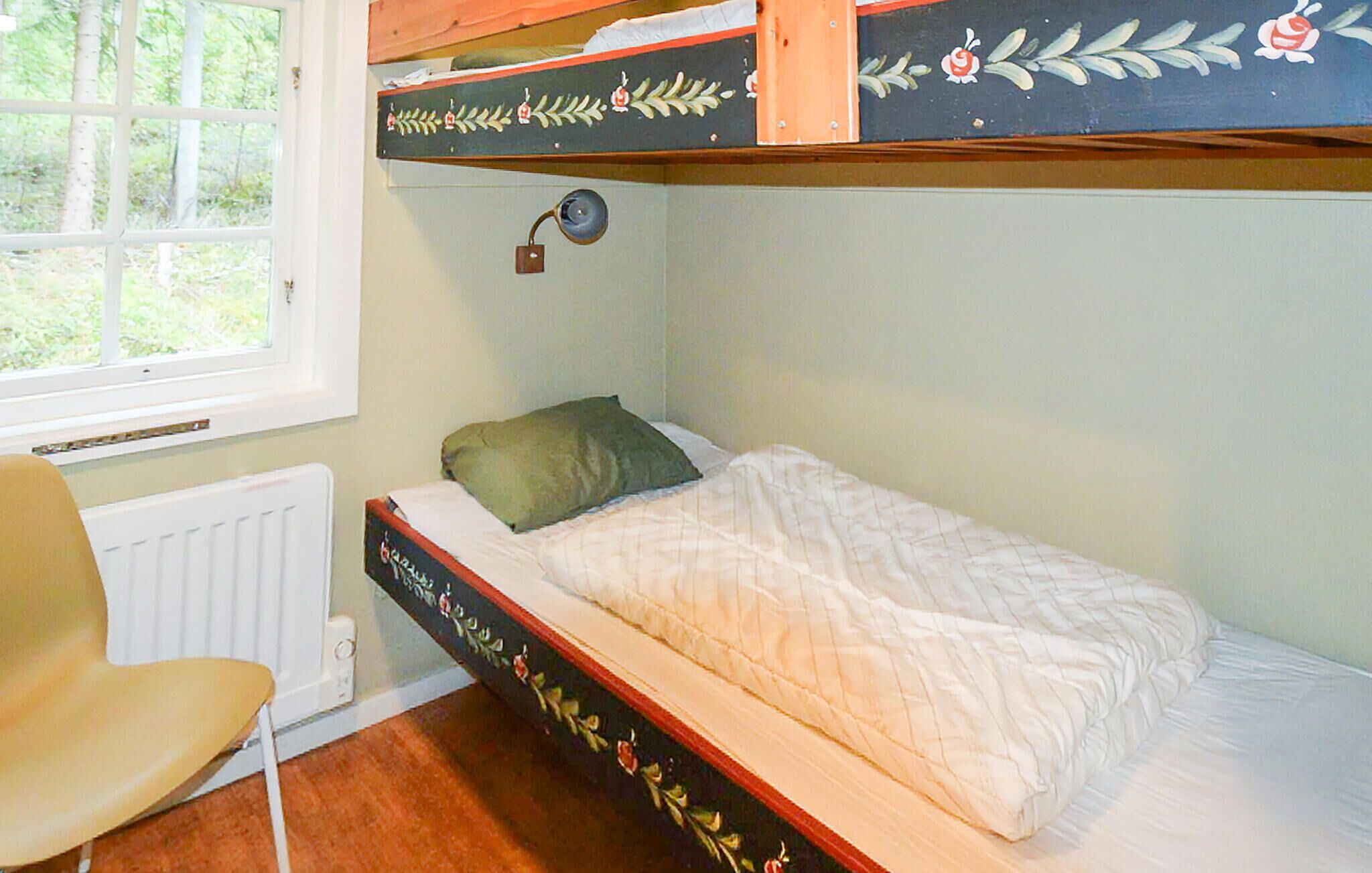 A bunk bed with a floral design, a single bed with a pillow, a chair, and a window with a view of trees.