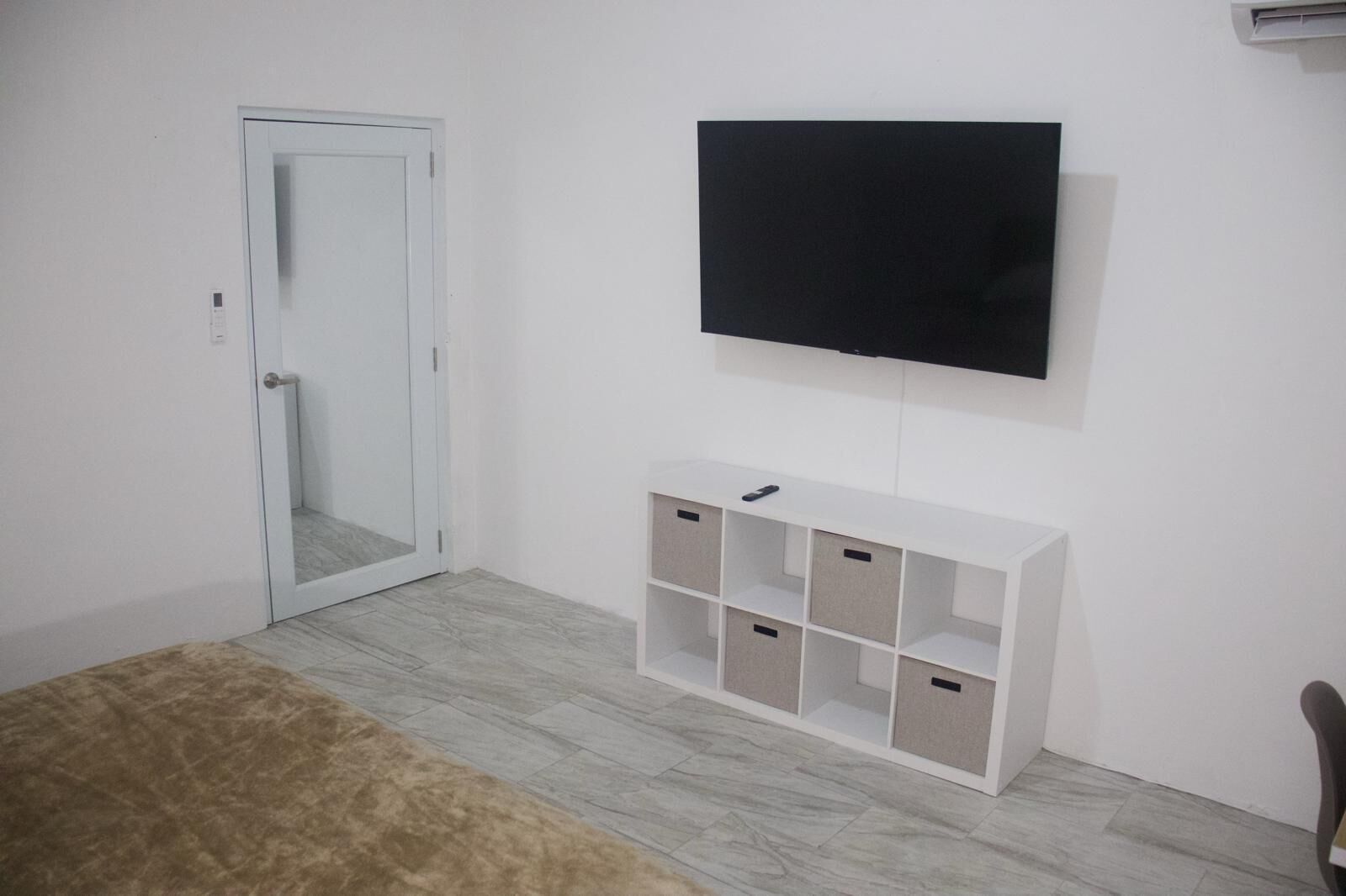 Una habitación con una puerta blanca, un televisor montado y un mueble blanco con varios compartimentos.