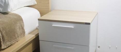 Ein kleiner Nachttisch mit Schubladen, ein Bett mit weißer Bettwäsche und ein Holzkopfstück.