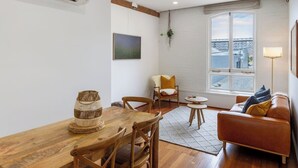 A cozy living room with a wooden dining table, chairs, a sofa, and a window with a view of a bridge. - New York Loft | Private Car Park | Walk to Stadium (Christchurch)