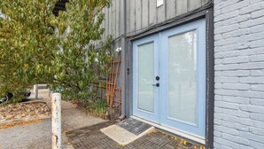 Una puerta azul con un ventanal, un muro de ladrillo blanco y un árbol de hojas verdes.