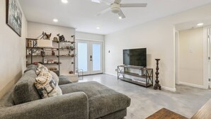 Una sala de estar con un sofá gris, un televisor sobre un mueble de madera, un ventilador de techo y una estantería con diversos objetos.