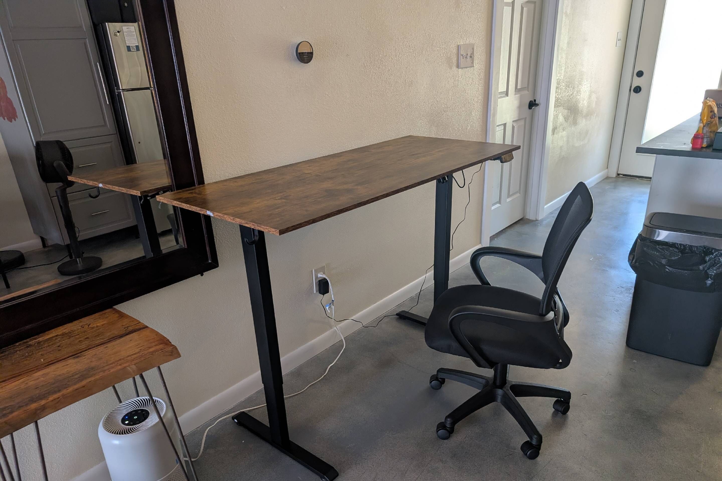 A room with a wooden desk, a black office chair, a white air purifier, a black trash bin, and a door leading to another room.