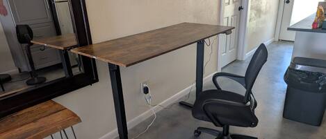 A room with a wooden desk, a black office chair, a white air purifier, a black trash bin, and a door leading to another room.