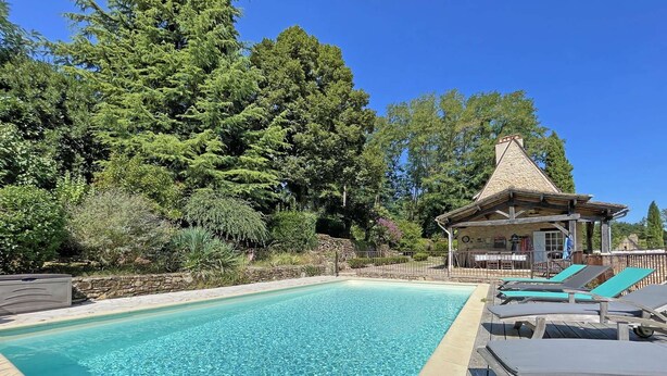 Una piscina cristallina con lettini e un pontile in legno, circondata da vegetazione lussureggiante e un edificio in pietra.