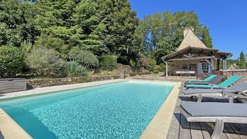 A clear swimming pool with lounge chairs and a wooden deck, surrounded by lush greenery and a stone building.