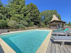 Une piscine aux eaux cristallines, dotée de chaises longues et d’une terrasse en bois, le tout entouré d’une végétation luxuriante et d’un bâtiment en pierre.